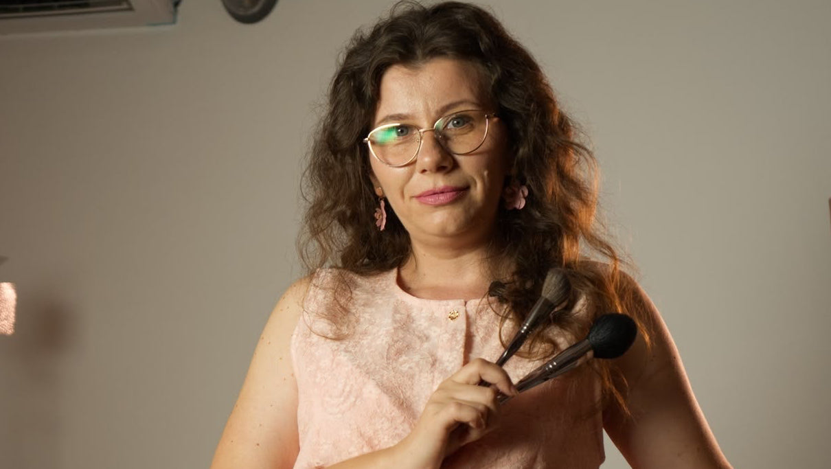 A woman with curly brown hair and glasses sits on a dark gray chair in a studio setting, holding a black handheld device. She is wearing a light peach-pink sleeveless jumpsuit.